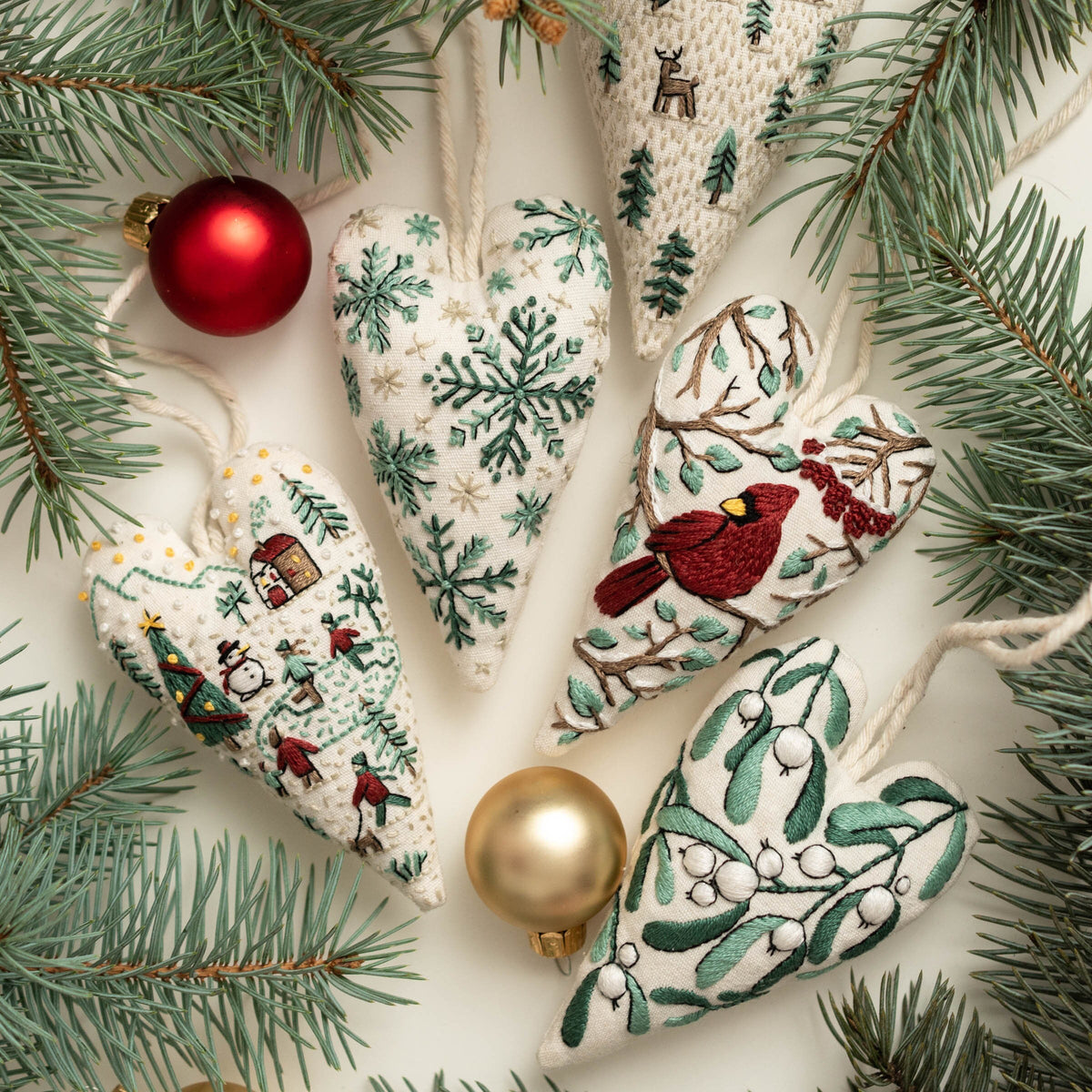 May include: Five handmade felt heart ornaments with embroidered winter scenes. The ornaments feature a cardinal, snowflakes, a winter village, and holly.