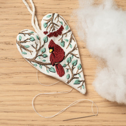 May include: A white heart-shaped fabric ornament with a red cardinal embroidered on a branch with green leaves. The ornament is hanging from a white string and there is a needle and thread on the wooden surface.