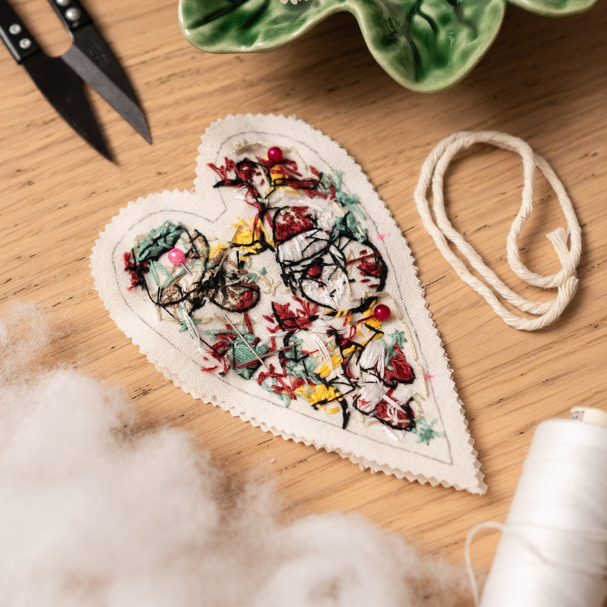 May include: A white heart-shaped fabric with a floral embroidery design in red, yellow, and green. The fabric is decorated with pink pins.