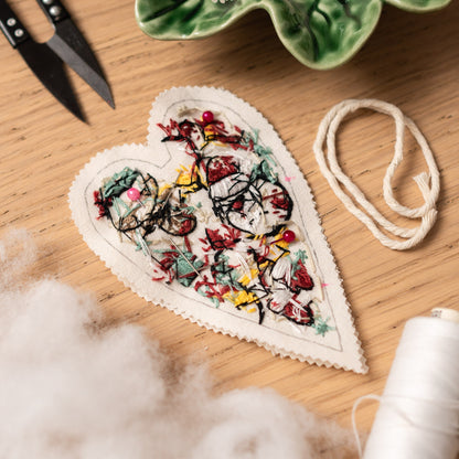 May include: A white heart-shaped fabric with a floral embroidery design in red, yellow, and green. The fabric is decorated with pink pins.