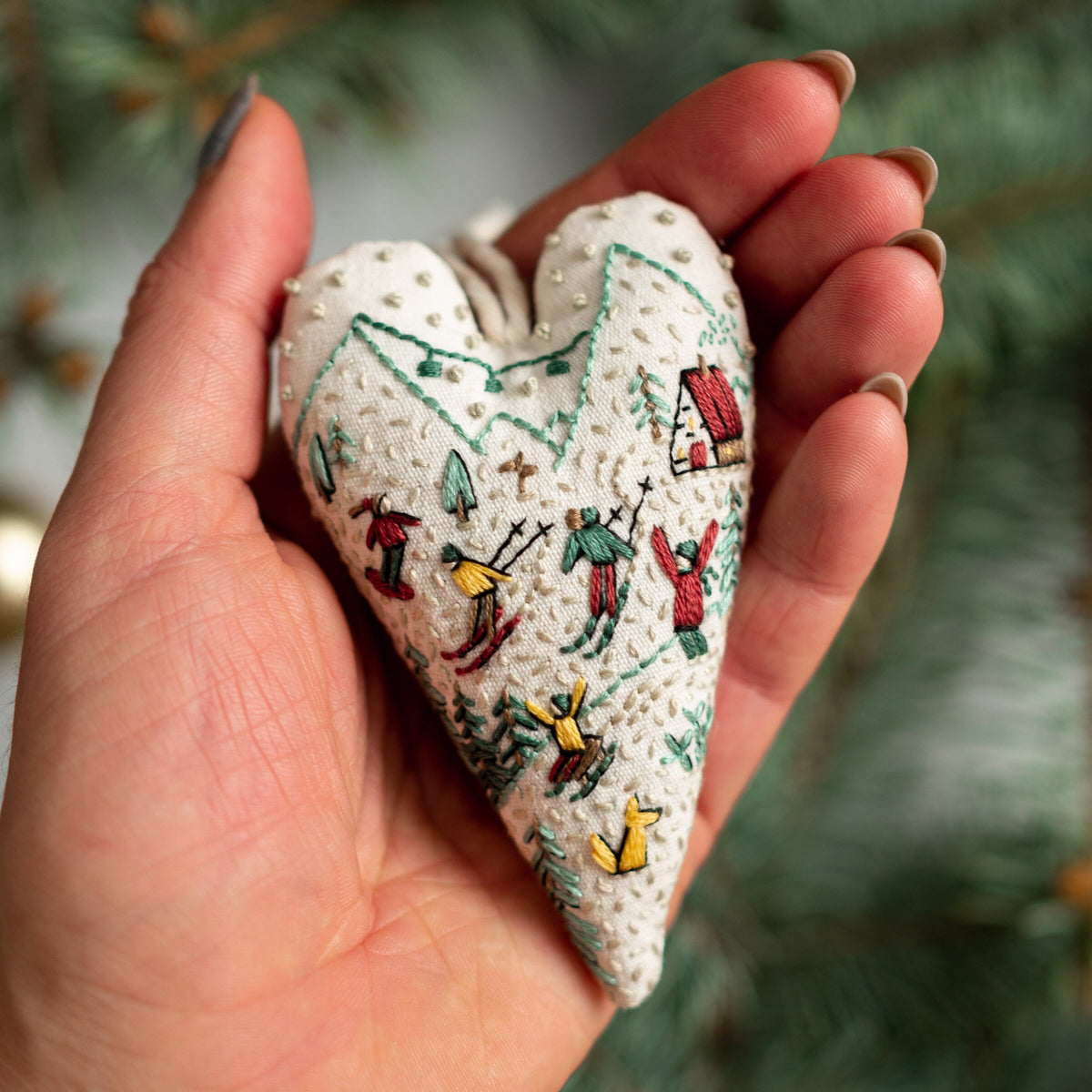 May include: A white heart-shaped fabric ornament with embroidered winter scenes. The ornament features skiers, a small house, and trees in green, red, and yellow thread.