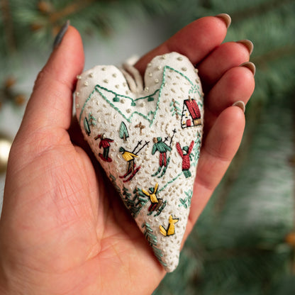 May include: A white heart-shaped fabric ornament with embroidered winter scenes. The ornament features skiers, a small house, and trees in green, red, and yellow thread.