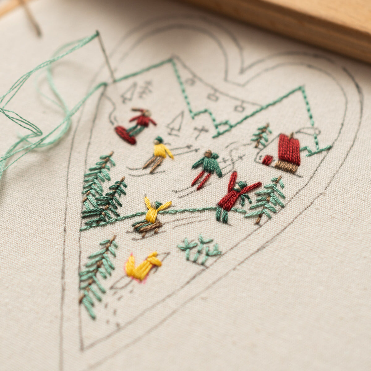 May include: Embroidered heart-shaped design on white fabric depicting a winter scene with skiers, trees, and a small cabin. The design is done in green, red, and yellow thread.