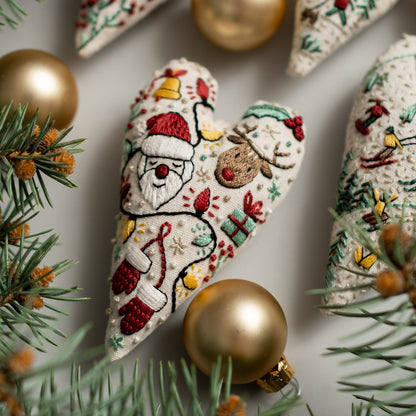 May include: A heart-shaped, cream-colored fabric ornament with embroidered Christmas scenes, including Santa Claus, a reindeer, and a string of lights. The ornament is surrounded by gold and green ornaments and a pine branch.