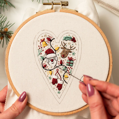May include: A close-up of a hand embroidering a heart-shaped design on a white fabric stretched in a wooden embroidery hoop. The design features a Santa Claus, a reindeer, a gift, and other Christmas-themed elements.