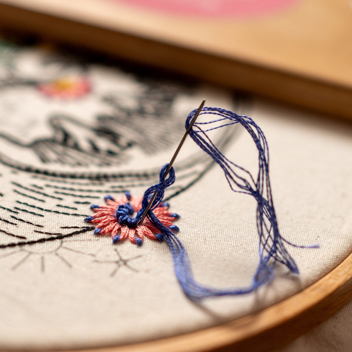 May include: Close-up of a needle and blue thread being used to embroider a pink and blue flower on a white fabric. The fabric is stretched over a wooden embroidery hoop.