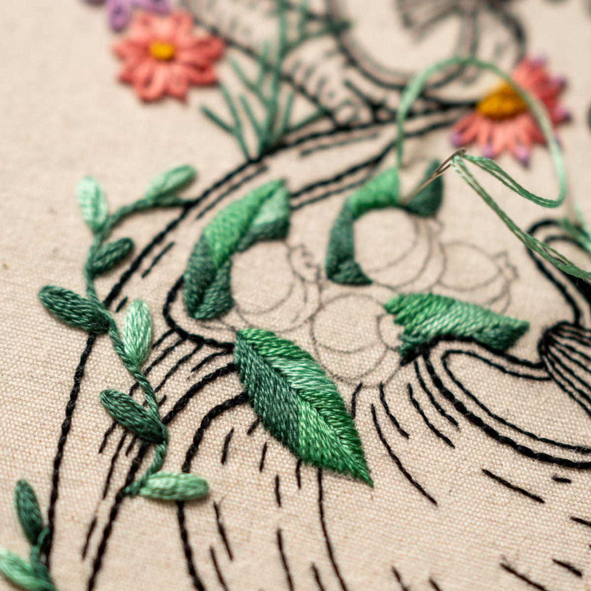 May include: Close-up of a green and black embroidery design on a white fabric. The design features leaves and vines, with a needle and thread visible in the background.