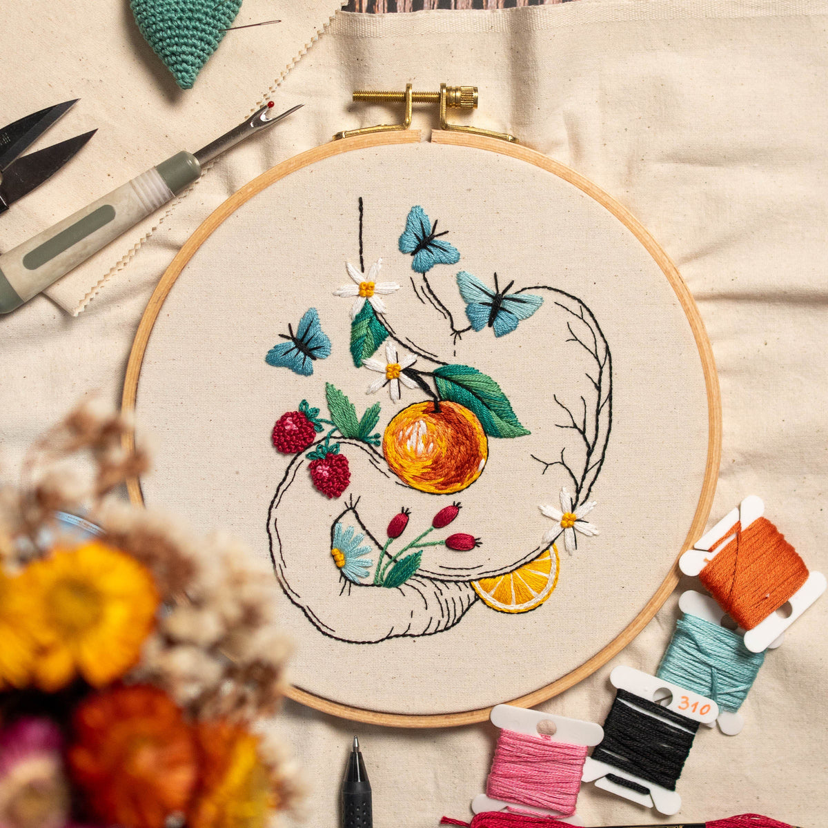 May include: An embroidery hoop with a botanical design featuring an orange, strawberries, butterflies, and daisies. The embroidery is done with colorful thread on a cream-colored fabric. Embroidery tools and spools of thread are visible.
