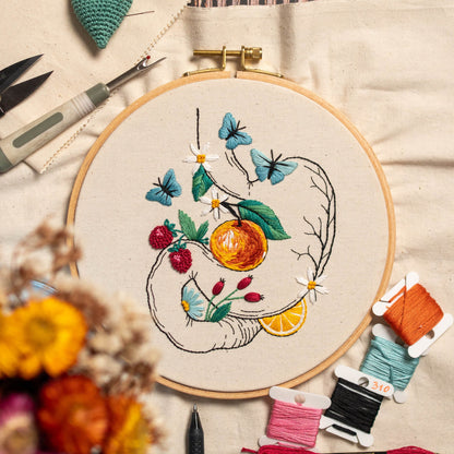 May include: An embroidery hoop with a botanical design featuring an orange, strawberries, butterflies, and daisies. The embroidery is done with colorful thread on a cream-colored fabric. Embroidery tools and spools of thread are visible.
