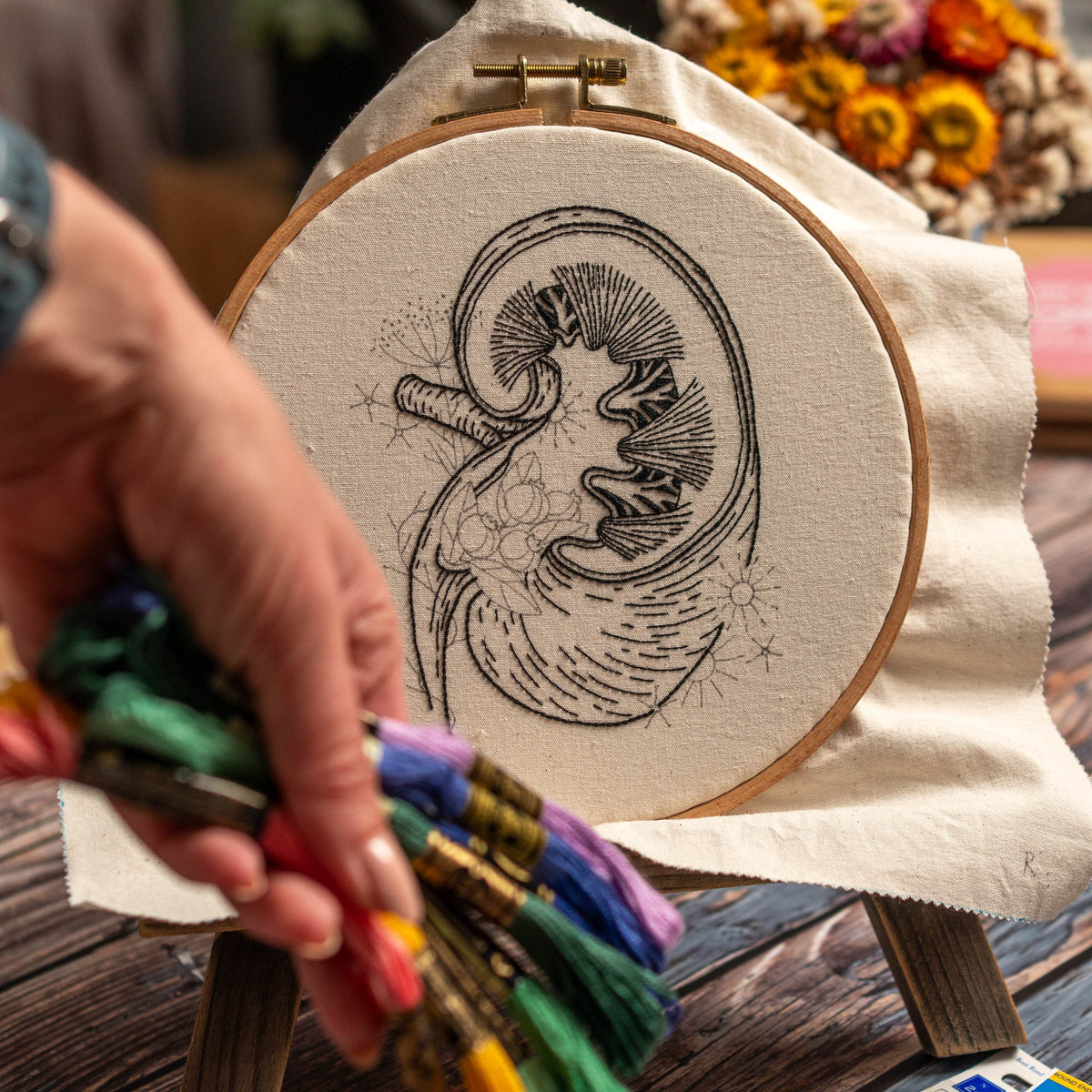 May include: A close-up of a hand holding embroidery floss next to a hoop with a black and white embroidery design of a kidney. The embroidery is on a white fabric.