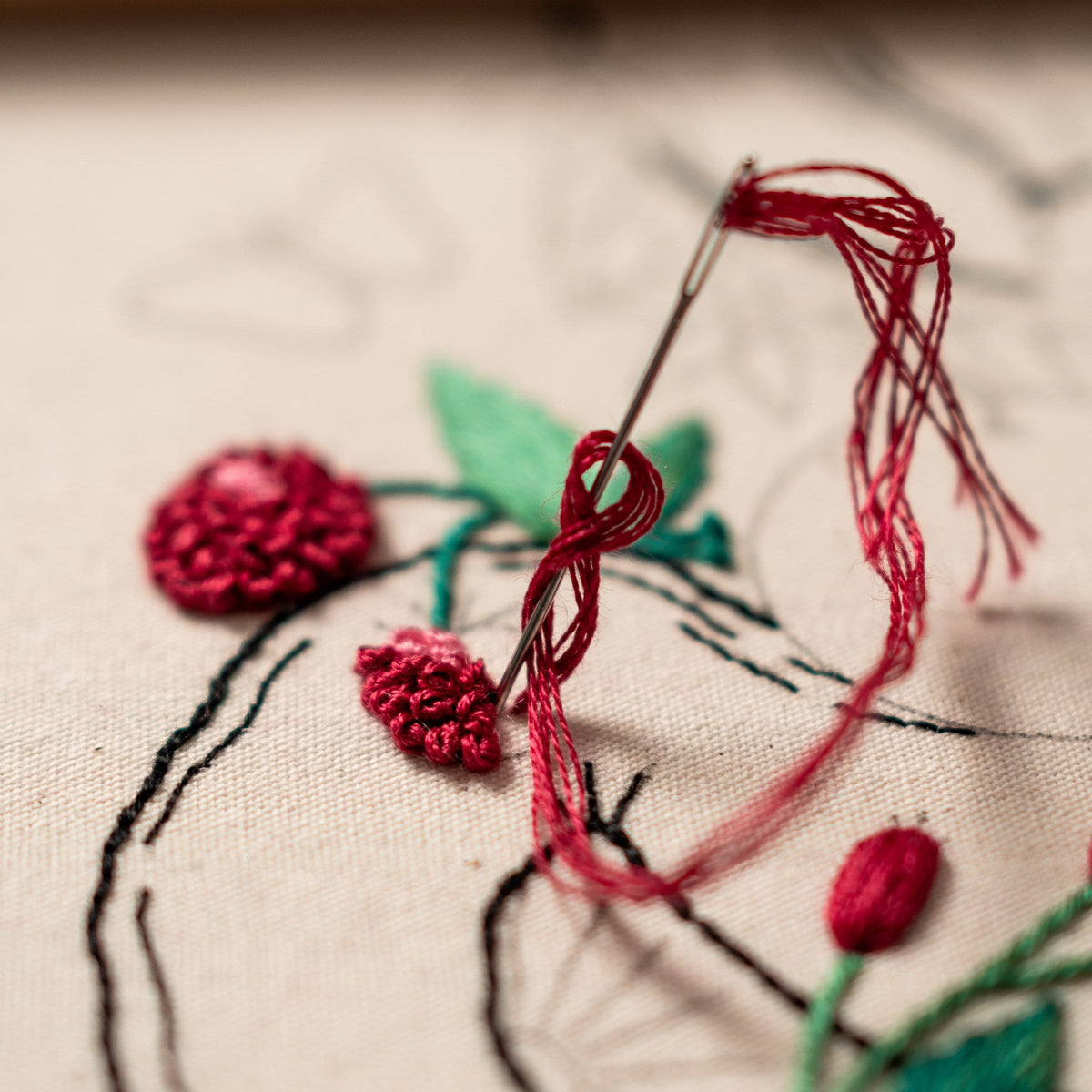 May include: Close-up of embroidery in progress. A needle threaded with red thread is piercing through the fabric. The fabric features embroidered red flowers and green leaves. The background is a light beige color.