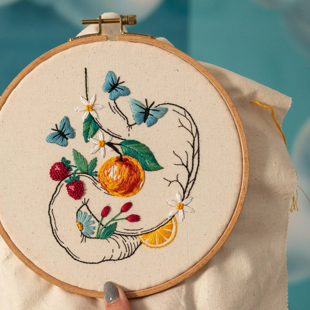 May include: Embroidered artwork in a wooden hoop. The design features a vibrant orange fruit, blue butterflies, red berries, and white flowers. The embroidery is on a cream-colored fabric, with a detailed, hand-stitched appearance.