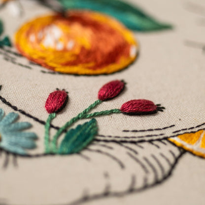 May include: Close-up of an embroidered design featuring a stylized orange fruit and red berries with green stems and leaves. The embroidery is on a light beige fabric, with black outline details.