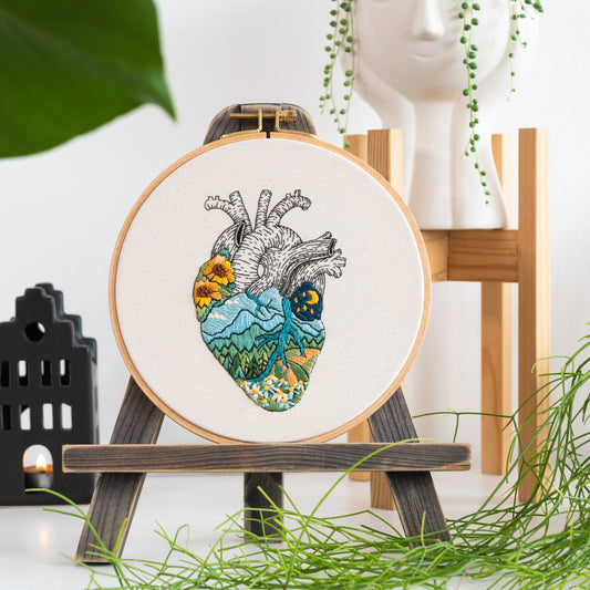 May include: Embroidered heart with a landscape design, including mountains, a river, and flowers. The heart is stitched on a white canvas and displayed on a wooden easel.