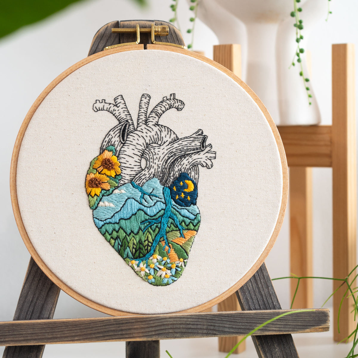 May include: Embroidered heart with a landscape design. The heart is filled with a mountain scene, flowers, and a night sky with a moon and stars. The embroidery is on a white canvas and is framed in a wooden hoop.