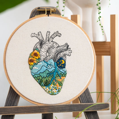 May include: Embroidered heart with a landscape design. The heart is filled with a mountain scene, flowers, and a night sky with a moon and stars. The embroidery is on a white canvas and is framed in a wooden hoop.
