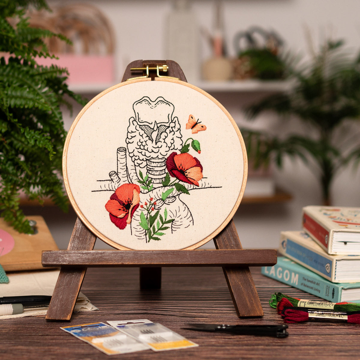 May include: Embroidered hoop art featuring a detailed anatomical illustration of the larynx with red poppy flowers and butterflies. The hoop is displayed on a wooden easel.
