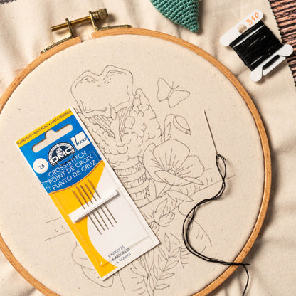 May include: A close-up of a cross-stitch embroidery hoop with a floral design. The hoop is made of wood and has a beige fabric stretched over it. A package of DMC cross-stitch needles is visible in the foreground. The needles are labeled "Cross-Stit