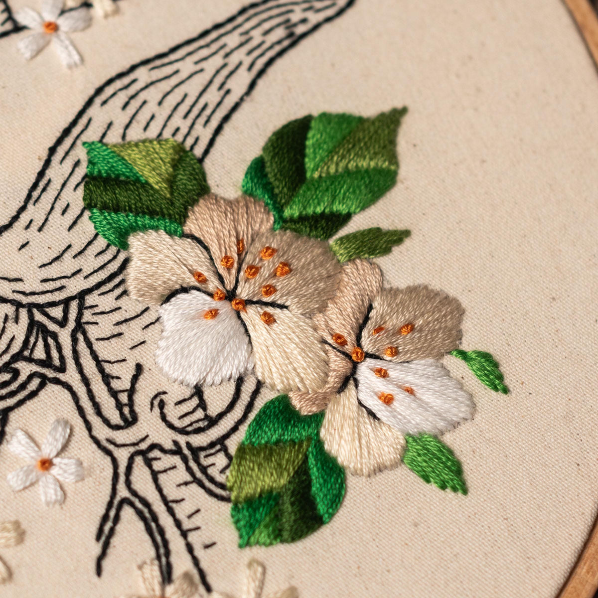 May include: Close-up of an embroidered design featuring white and beige flowers with orange centers, green leaves, and a black and white outline of a branch.