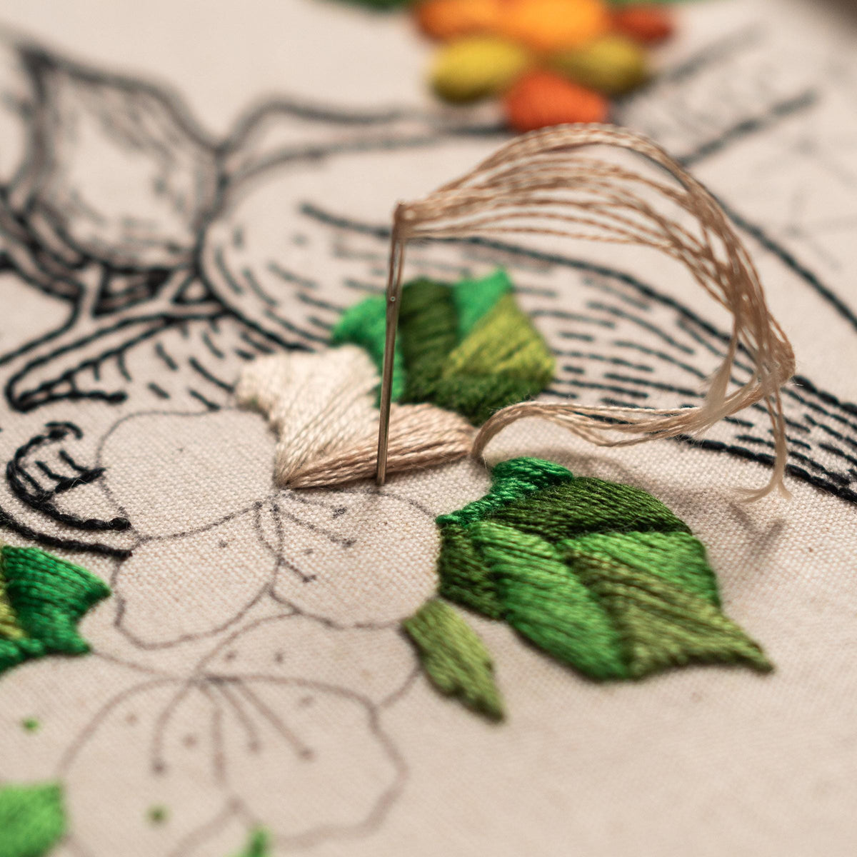 May include: Close-up of a needle and thread being used to embroider a flower design on a piece of fabric. The flower is white with green leaves and is being stitched with a beige thread.
