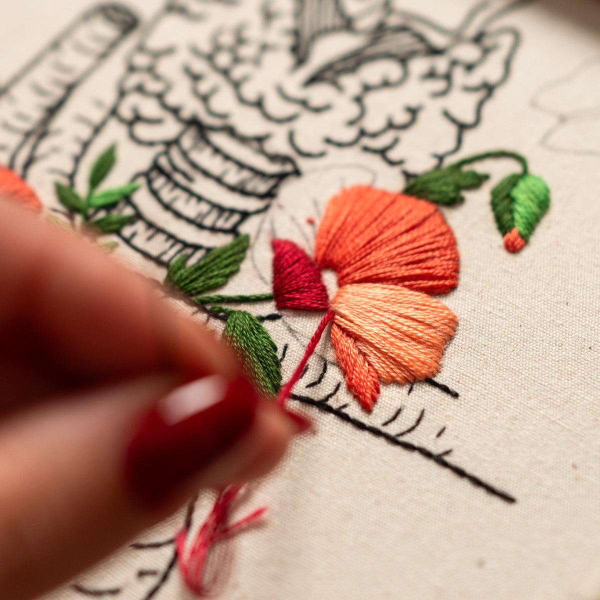 May include: Close-up of a hand embroidering a flower with orange and red thread on a white fabric. The flower is surrounded by green leaves and a black outline.