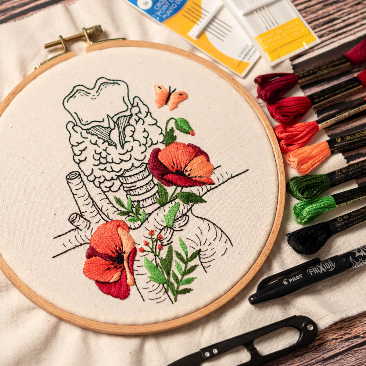 May include: Embroidered hoop art featuring a detailed anatomical illustration of the human trachea with red poppy flowers and green leaves. The hoop is made of wood and has a white fabric background.