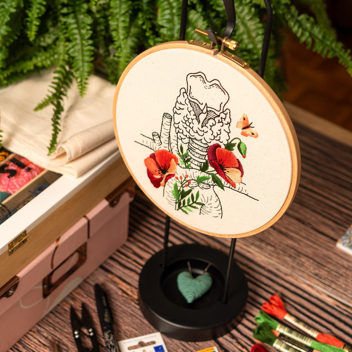 May include: A white embroidery hoop with a black and white anatomical illustration of the larynx and trachea with red poppies and a butterfly. The hoop is on a black stand with a green heart.