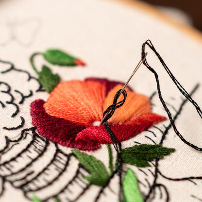 May include: Close-up of a needle and thread being used to embroider a red and orange flower on a white fabric. The flower is surrounded by green leaves.