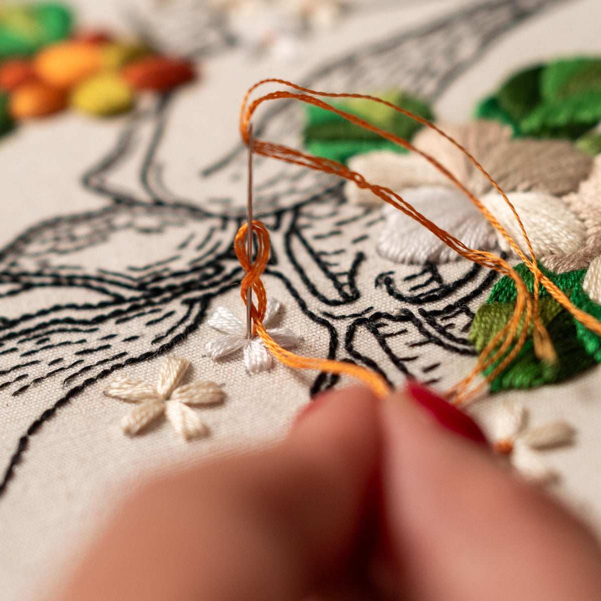 May include: Close-up of a hand embroidering a white flower with orange thread on a white fabric. The fabric has a black line drawing of a landscape with other embroidered flowers.