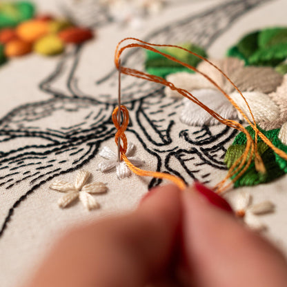 May include: Close-up of a hand embroidering a white flower with orange thread on a white fabric. The fabric has a black line drawing of a landscape with other embroidered flowers.