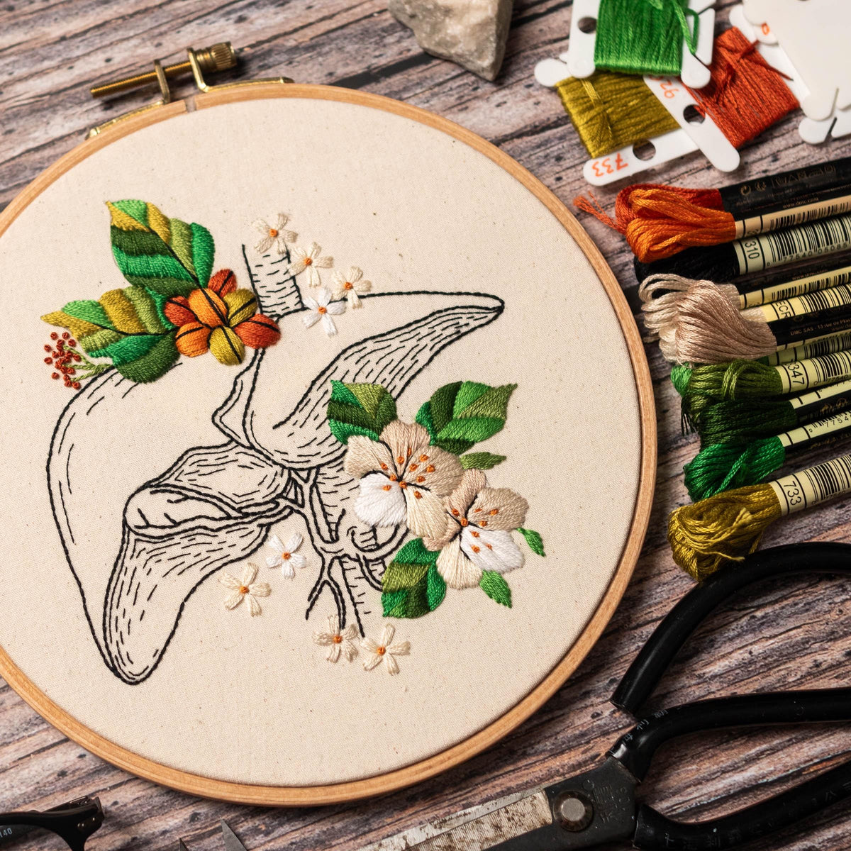 May include: Embroidered hoop art featuring a detailed anatomical illustration of a liver with delicate floral embellishments. The design includes white flowers, green leaves, and orange accents.