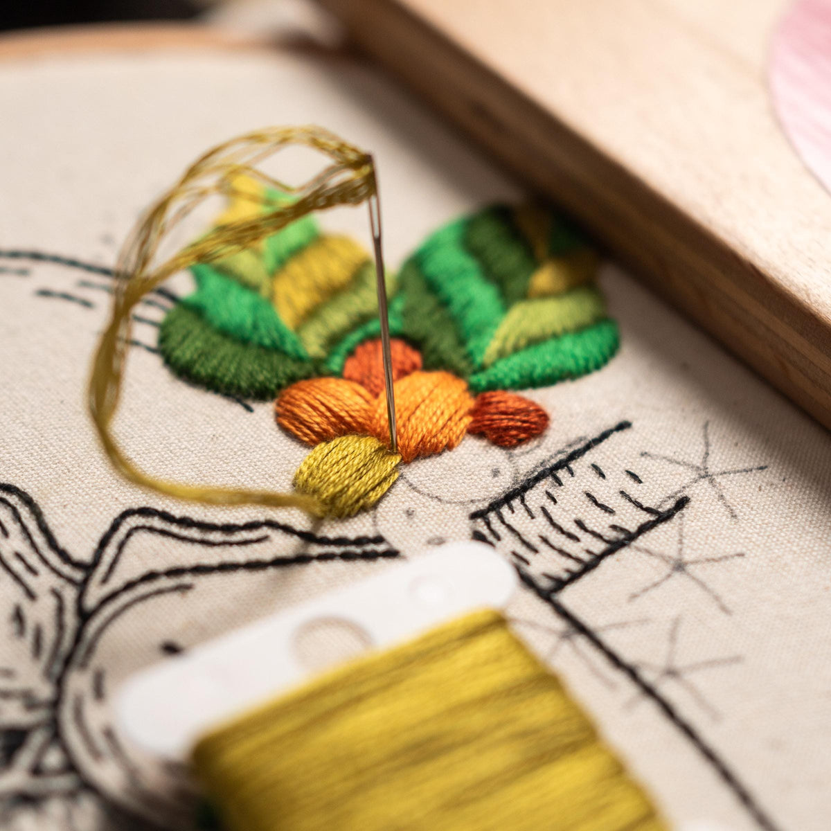 May include: Close-up of a needle and thread being used to embroider a green, yellow, and orange flower on a white fabric. The fabric has a black and white design printed on it.