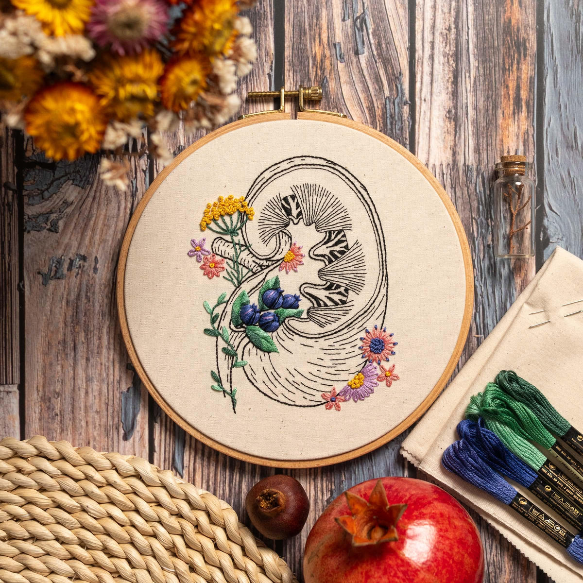 May include: Embroidered hoop art featuring a detailed anatomical illustration of a kidney with delicate floral accents in shades of pink, yellow, and blue. The hoop is made of wood and has a natural linen fabric.