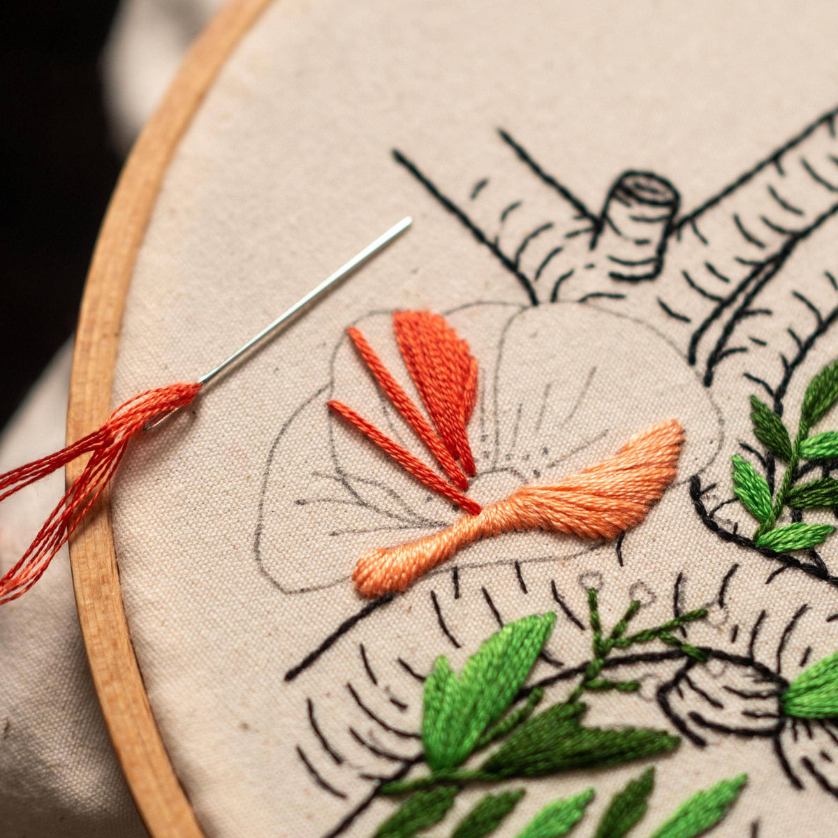 May include: Close-up of a hand embroidery project in progress. A floral design with orange and green thread is stitched on a white fabric stretched in a wooden embroidery hoop. A needle with orange thread is visible.