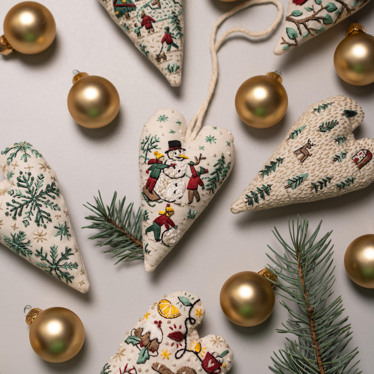 May include: A collection of six handmade heart-shaped ornaments with embroidered winter scenes. The ornaments are made of white fabric with green, red, and brown embroidery. The ornaments are decorated with gold beads and are surrounded by gold orna