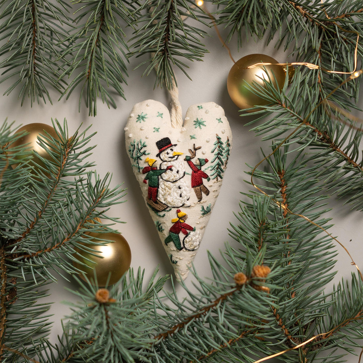May include: A white heart-shaped ornament with an embroidered scene of children playing in the snow with a snowman. The ornament is surrounded by green pine branches and gold Christmas lights.