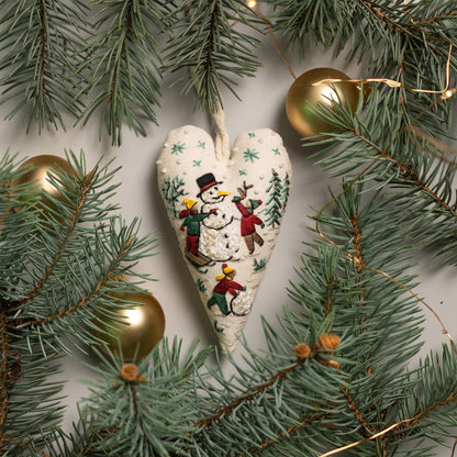 May include: A white heart-shaped ornament with an embroidered scene of children playing in the snow with a snowman. The ornament is surrounded by green pine branches and gold Christmas lights.