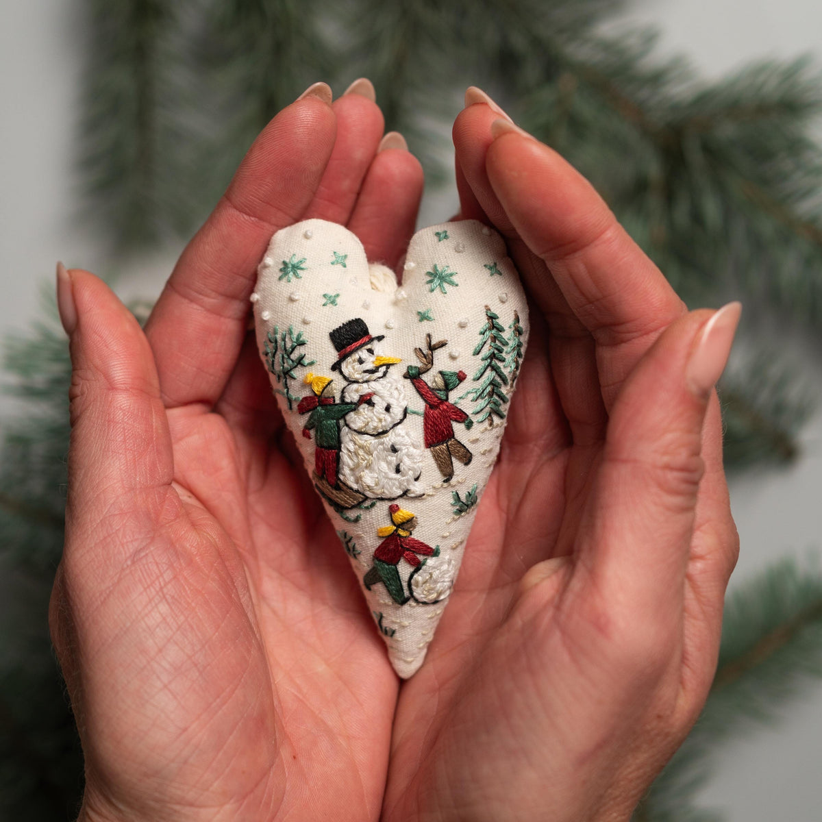 May include: A white heart-shaped fabric ornament with embroidered snowflakes and a winter scene of a snowman and children playing.