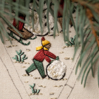 May include: Embroidered image of a person in a red and green outfit rolling a snowball in a snowy landscape. The person is wearing a yellow hat and a red scarf. The background is a white canvas with green embroidery details.