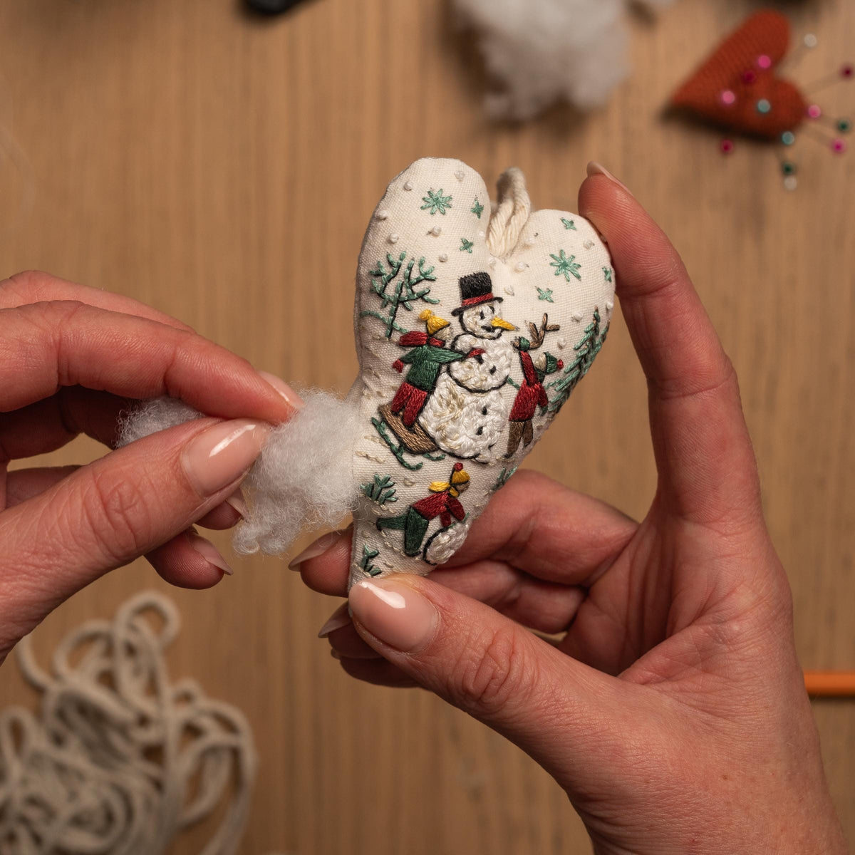 May include: A white heart-shaped fabric ornament with an embroidered scene of a snowman and children playing in the snow. The ornament is being stuffed with white fiberfill.