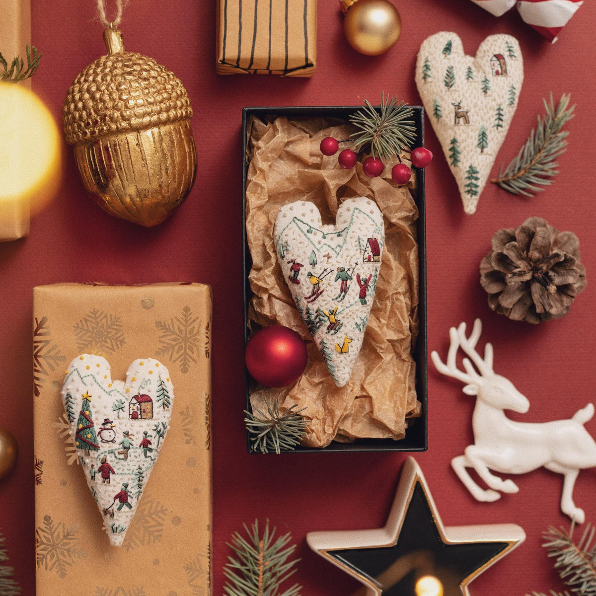 May include: A set of three handmade felt heart ornaments with winter scenes embroidered on them. The ornaments are hanging from a red ribbon and are surrounded by other Christmas decorations.