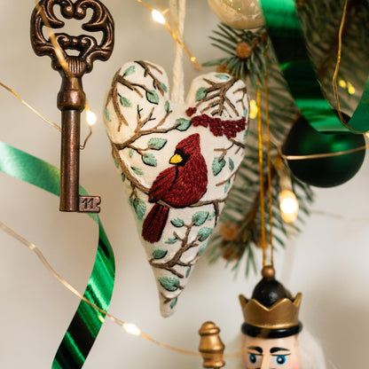 May include: A white heart-shaped ornament with red and green embroidery depicting a cardinal bird perched on a branch. The ornament is hanging from a string and is surrounded by Christmas lights and greenery.