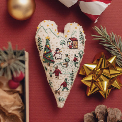 May include: A white heart-shaped fabric ornament with an embroidered winter scene. The scene includes a snowman, a Christmas tree, a house, and people ice skating. The ornament is perfect for adding a festive touch to your home decor.