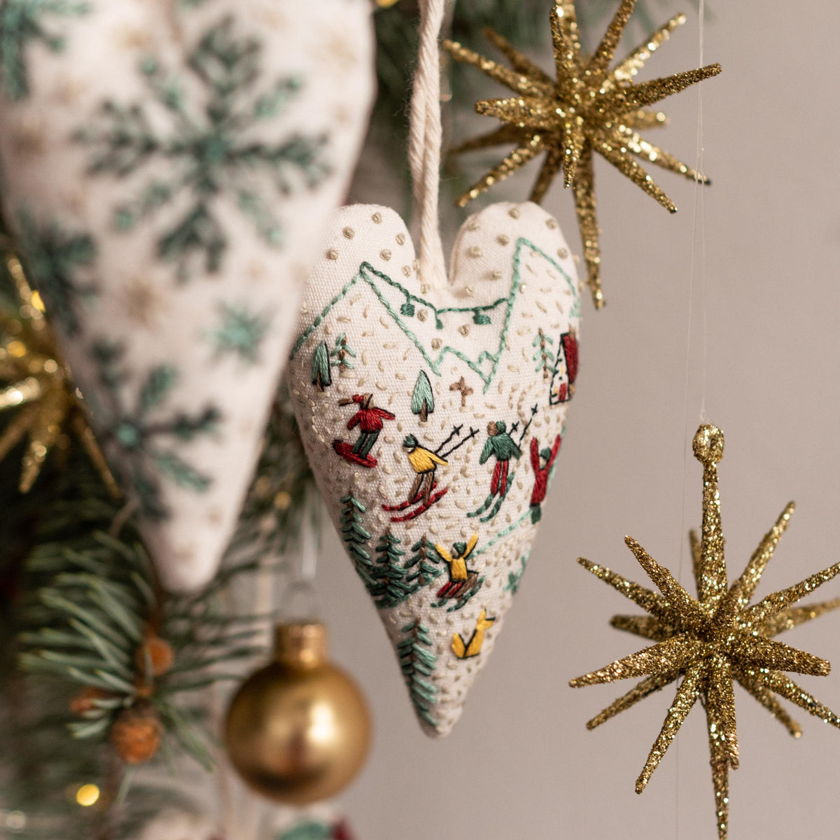 May include: White felt heart-shaped ornament with an embroidered scene of skiers and mountains. The ornament is hanging on a Christmas tree with gold star ornaments.