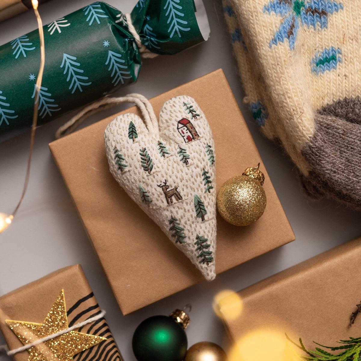 May include: A white heart-shaped ornament with a brown house, a deer, and green trees embroidered on it. The ornament is sitting on a brown paper gift bag with a gold ornament and a green ornament nearby.