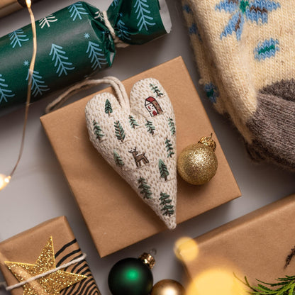 May include: A white heart-shaped ornament with a brown house, a deer, and green trees embroidered on it. The ornament is sitting on a brown paper gift bag with a gold ornament and a green ornament nearby.