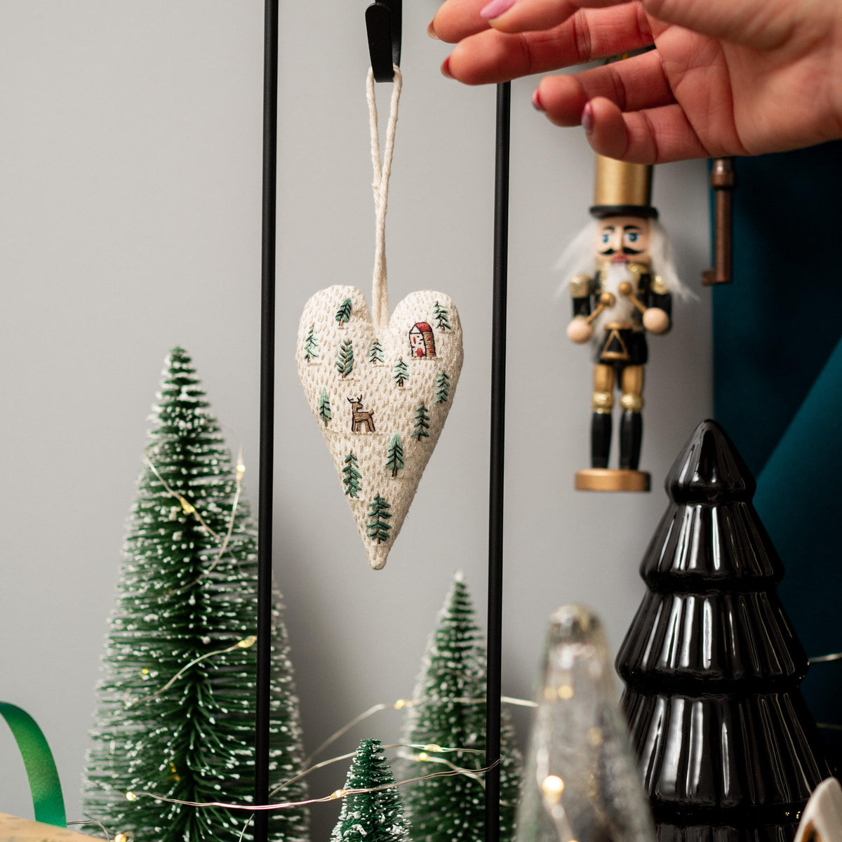 May include: A white heart-shaped ornament with a winter scene embroidered on it. The scene includes a small house, trees, and a deer. The ornament is hanging from a string.