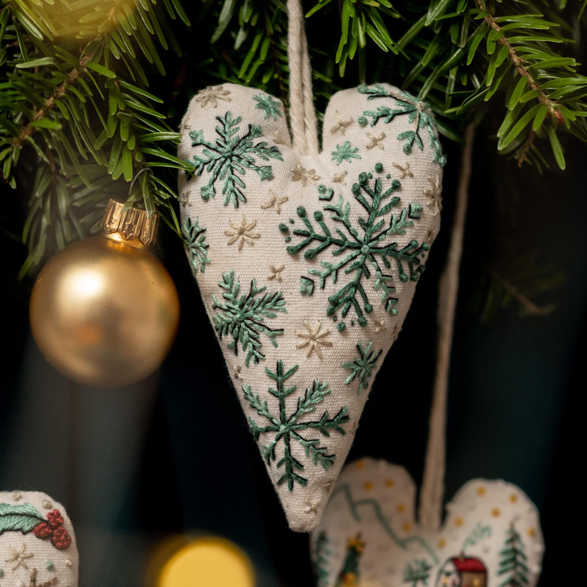 May include: A white heart-shaped ornament with green snowflake embroidery hanging from a Christmas tree. The ornament is made of fabric and has a rustic, handmade look.