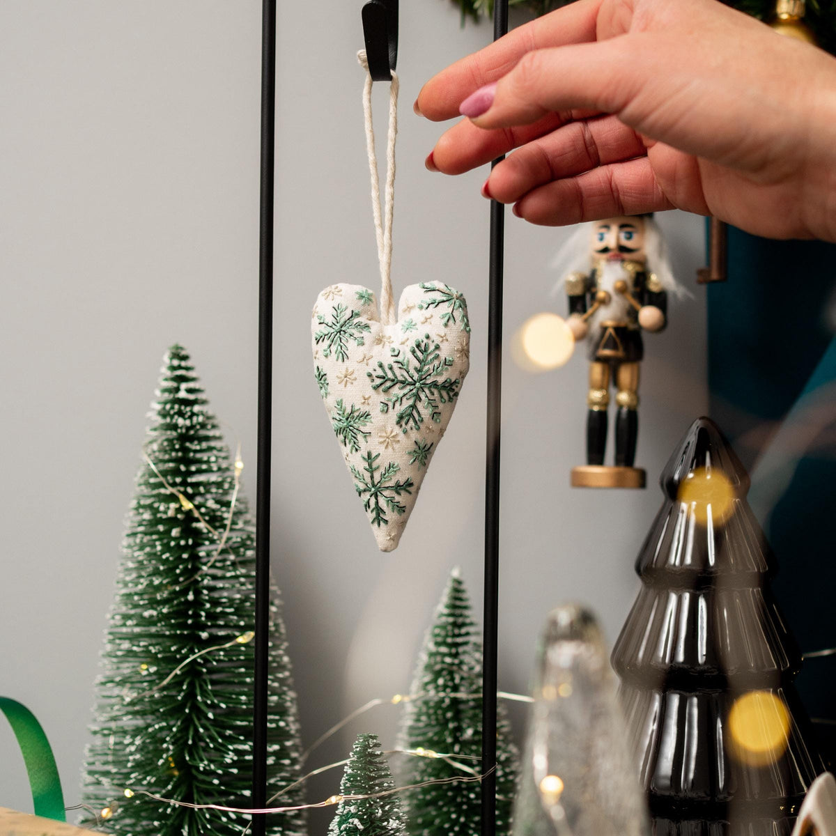 May include: A white heart-shaped ornament with green snowflake embroidery hangs from a string. The ornament is in front of a black metal stand and a small green Christmas tree.