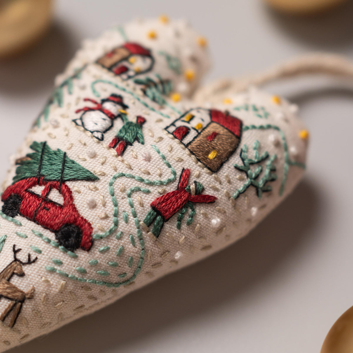 May include: A heart-shaped ornament with a winter scene embroidered on it. The scene includes a car with a Christmas tree on top, a snowman, a house, and a person carrying a tree. The ornament is made of white fabric and has a loop for hanging.